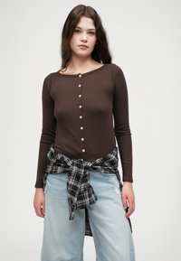 Haut marron côtelé à manches longues avec encolure ronde et boutons sur le devant, associé à un jean large déchiré bleu clair et une chemise à carreaux noire nouée à la taille.