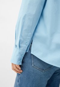 Lichtblauw langemouwen shirt met knoopmanchetten, met een gladde textuur, zijsplitten en een relaxed fit, gecombineerd met spijkerbroek.