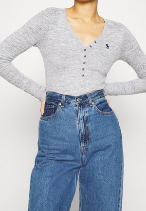 Personne portant une chemise henley grise à manches longues avec des boutons métalliques et un jean bleu taille haute, les mains reposant sur les hanches.