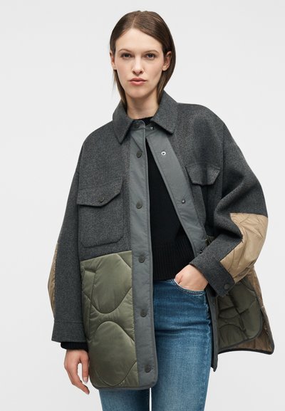 Γυναίκα που φοράει γκρι και ελιάς παλτό με καπιτονέ πατς πάνω από μαύρο πουλόβερ και μπλε τζιν, με το ένα χέρι στην τσέπη της, με ουδέτερο φόντο.