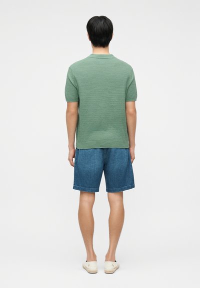 Άνδρας που στέκεται και κοιτάζει προς τα πίσω, φορώντας πράσινη πλεκτή πόλο με κοντά μανίκια, μπλε τζιν σορτς και άσπρα παπούτσια χωρίς κορδόνια.