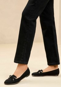 Des ballerines en suede noir avec un bout rond, un accent en nœud et des accessoires en métal argenté, le tout reposant sur une fine semelle blanche. Portées avec un jean noir.