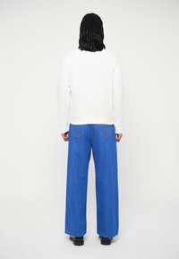 Sweatshirt branco com punhos e barra canelados, combinado com jeans azul de perna larga, apresentando costuras subtis nos bolsos traseiros. Sapatos pretos.