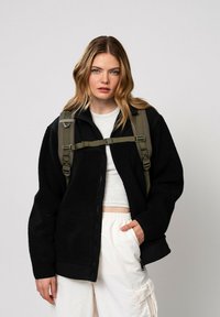 Veste en polaire noire avec fermeture éclair et poignets élastiques, associée à un sac à dos vert olive et un pantalon cargo blanc. Texture douce et coupe décontractée.