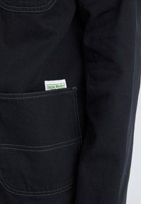 Black work jacket featuring a front pocket with white stitching and a green label that reads "Service Works." Durable fabric texture.