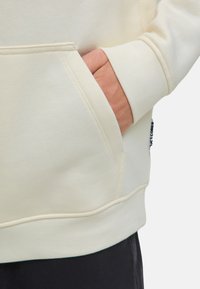 Hoodie beige clair avec une poche avant, montrant une main reposant dans la poche, associé à un pantalon foncé. Texture de tissu douce visible.