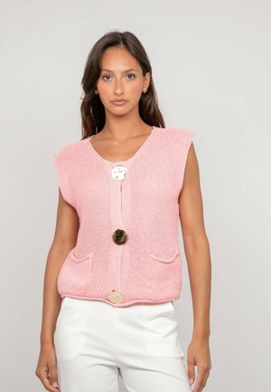 Femme portant un gilet en tricot rose sans manches avec trois grands boutons et deux poches avant, associée à un pantalon blanc, debout devant un fond uni.