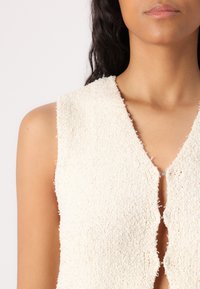 Haut sans manches texturé blanc avec un décolleté en V et un design à fermeture ouverte, présentant des bords bruts et un tissu duveteux. Pas de matériel visible.