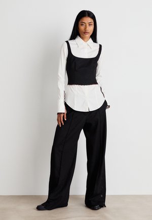 Haut noir à manches larges et courtes sur une chemise blanche à boutons. Un pantalon noir ample présente un motif subtil et des accents de boutons rouges.