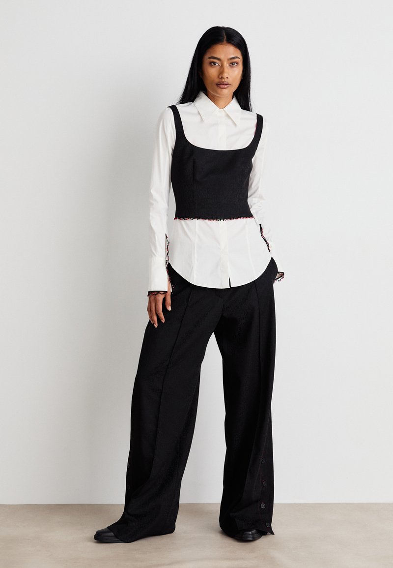 Haut noir à manches larges et courtes sur une chemise blanche à boutons. Un pantalon noir ample présente un motif subtil et des accents de boutons rouges.