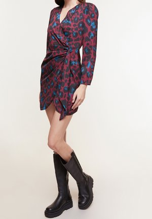 Femme portant une robe portefeuille imprimé animal bordeaux et bleu à manches longues avec des bottes noires épaisses montant jusqu'aux genoux.