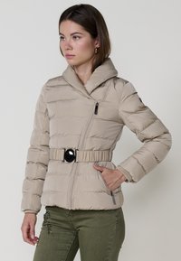 Bežna puf jakna s visokim ovratnikom, zatvorenim prednjim dijelom i sužavajućim strukom koji ima crnu kopču. Ima quilted teksturu i bočne džepove.