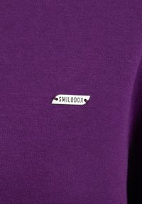 Una tela de punto morada con una textura suave presenta una pequeña etiqueta plateada etiquetada como "SMILODOX" cosida en el lateral.
