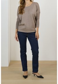 Kobieta ubrana w taupe'owy sweter z długim rękawem, ciemnogranatowe dżinsy i czarne buty na niskim obcasie w kształcie stożka stojąca na drewnianej podłodze.