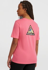Lyserød t-shirt lavet af bomuld, med et stort sort trekantet grafisk motiv på ryggen, der har farverige designelementer og brandtekst.