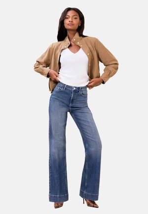 Naine, kes kannab liivavärvi nööbitud jopet, valget varrukateta topi, kõrge vöökohaga laiade jalgadega siniseid teksaseid ja konnahalliga kõrgete kontsadega kingi.