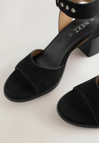 Two black suede high-heeled sandals with open toes and ankle straps, displayed against a neutral background.