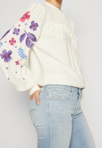 Vitt blus med broderade blommönster i lila, rosa och blått på puffärmar, kombinerat med ljusblå jeans. Har knappar.