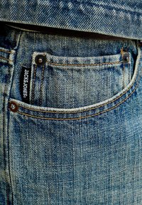 Nærbillede af lomme på blå denimjeans med synlige syninger, metalnitter og en sort stofmærkat med hvid tekst.