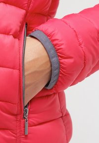 Veste matelassée rose avec un design matelassé, dotée d'un poignet gris et d'une poche zippée latérale. Le tissu semble lisse et isolant.