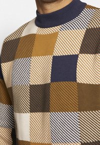Pull en tricot avec un motif à carreaux géométrique dans des tons de marron, bleu marine, crème et moutarde. Col bleu marine côtelé. Tissu doux et texturé.