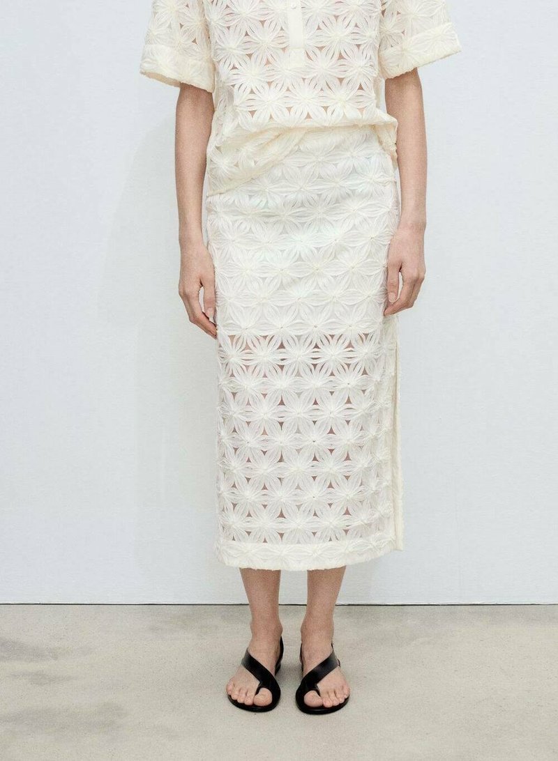 Ensemble en top et jupe transparents blancs avec un motif floral. Le tissu présente des broderies délicates et une silhouette droite. Sandales noires.