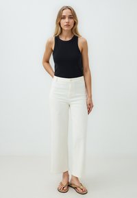 Top senza maniche nero, jeans a gamba larga bianco sporco con tasche frontali e infradito marroni con dettaglio decorativo della fibbia.