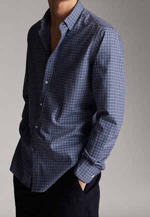 Homme portant une chemise à carreaux bleue à manches longues avec des boutons, partiellement rentrée dans un pantalon noir, debout avec une main dans la poche.