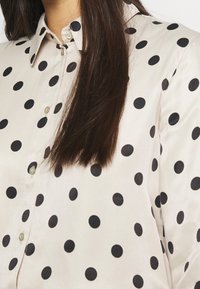 Camicia beige con bottoni realizzata in tessuto morbido, decorata con grandi pois neri, dotata di un colletto classico e dettagli di chiusura con bottoni discreti.