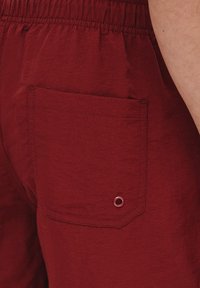 Bordowe szorty z tylną kieszenią z prostokątną klapką i małym metalowym oczkiem. Materiał ma teksturowany wygląd.