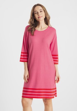 Vrouw die een loszittende roze jurk draagt met driekwart mouwen en rode horizontale strepen op de mouwen en zoom, staand tegen een witte achtergrond.