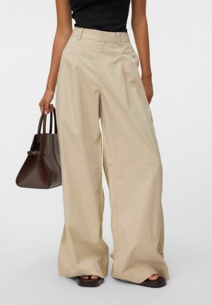 Femme portant un pantalon beige taille haute à jambes larges, un top noir sans manches, portant un sac à main marron foncé et des sandales ouvertes foncées.