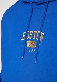 Blå bomullshuvtröja med dragsnören, prydd med guldbrodyr som står "BOSTON USA 1987." Slät textur, medelhårdhet.