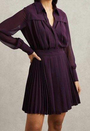 Vrouw die een donkerpaarse jurk met lange mouwen draagt, geplooid, met een nauwsluitende taille en een kraag