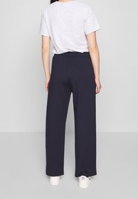 Persoon met de rug naar de camera, gekleed in een wit T-shirt, donkere wijde broek en witte Adidas sneakers, op een lichtgrijze vloer.