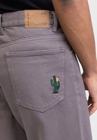 Pessoa vestindo calças cinzas com um bolso traseiro que exibe um pequeno cacto verde e um bordado de sol laranja, além de um patch de marca em couro castanho.