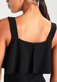 Czarny top bez rękawów z warstwowym projektem falbanek z tyłu oraz ukrytym zamkiem błyskawicznym. Materiał wydaje się teksturowany i matowy.