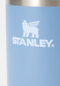 Niebieski kubek stalowy z matowym wykończeniem. Posiada biały logo korony misia z skrzydłami. Gładka tekstura, cylindryczny kształt.