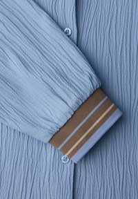 Světle modrá texturovaná látka košile s manžetou v hnědé, modré a béžové pruhované barvě. Má zapínání na knoflíky a zvlněný povrchový design.