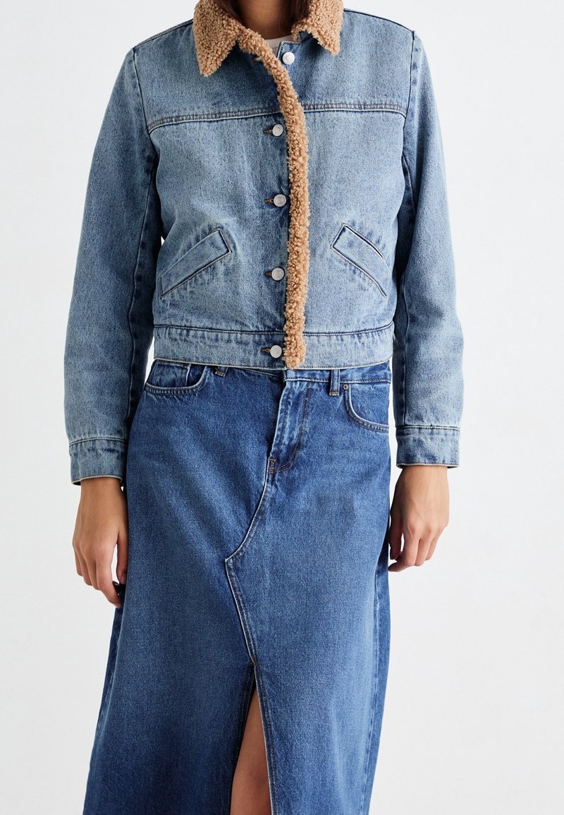 Femme portant une veste en jean bleu clair avec doublure polaire beige et une jupe en jean bleue avec une fente à l'avant.