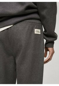 Pantalons de survêtement gris avec une taille élastique, arborant une petite étiquette sur la poche arrière portant la mention "Les lundis sont optionnels." Tissu doux et texturé.