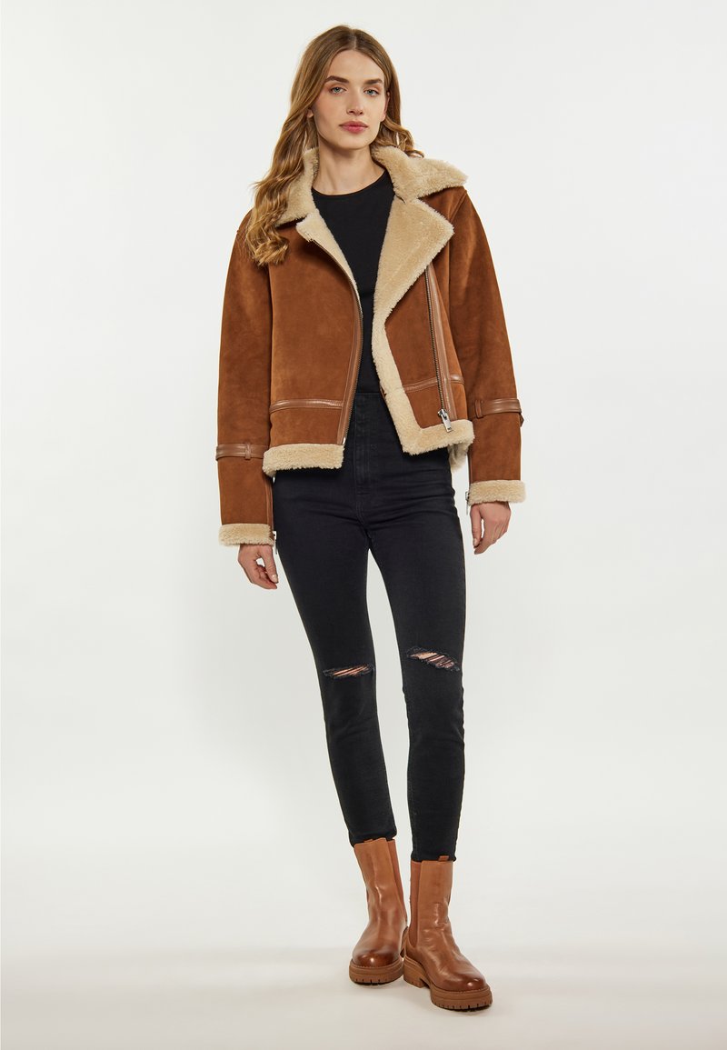 Brun shearlingjakke med beige foring, lynlåslukning og sidelommer; parret med sorte slidte jeans og brune ankelstøvler med tykke såler.