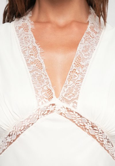 Witte kleding met fijne kantafwerking langs een diepe V-halslijn en het lichaam, die een gladde textuur met subtiele rafelaccenten toont.