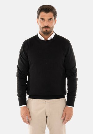 Maglione nero a girocollo realizzato in tessuto testurizzato. Maniche lunghe con polsini a costine, indossato sopra una camicia bianca con colletto. Pantaloni beige visibili.