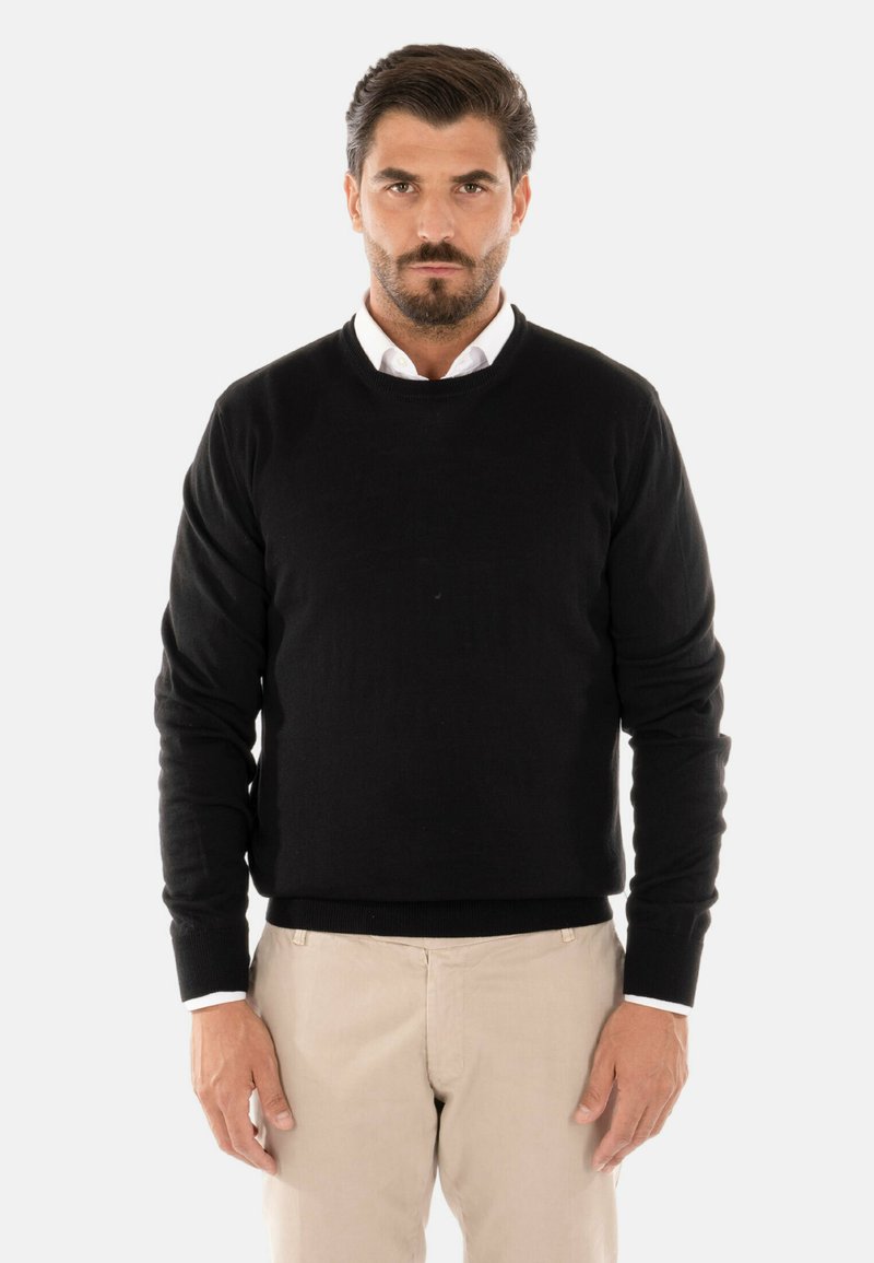 Maglione nero a girocollo realizzato in tessuto testurizzato. Maniche lunghe con polsini a costine, indossato sopra una camicia bianca con colletto. Pantaloni beige visibili.
