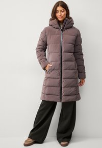 Abrigo largo acolchado en tono malva apagado con capucha, que presenta secciones acolchadas verticales y un cierre frontal. Combinado con pantalones de pierna ancha negros y zapatos.