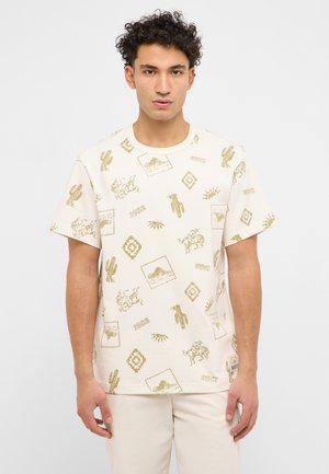 Young man wearing cream t-shirt and pants; t-shirt has gold western-style prints of cacti, horses, sun, and geometric shapes.