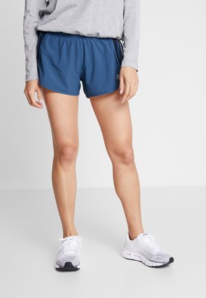 Person iført blå atletikshorts, grå langærmet skjorte og hvide løbesko, står på en grå gulv.