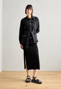 Černá džínová bunda s páskem v pase, dvěma náprsními kapsami a stříbrnými knoflíky, v kombinaci s úzkou černou žebrovanou sukní a robustními černými sandály.