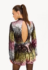 Jumpsuit pailleté avec des manches longues, dos ouvert, et couleurs multicolores de violet, doré et argenté. Texture lisse, design ajusté.